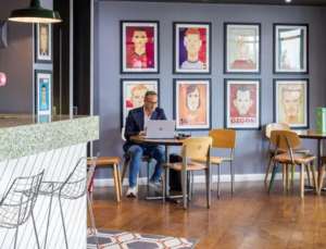 A man works at a table on his laptop in a coffee shop. Behind him are stylised portraits of sports players.